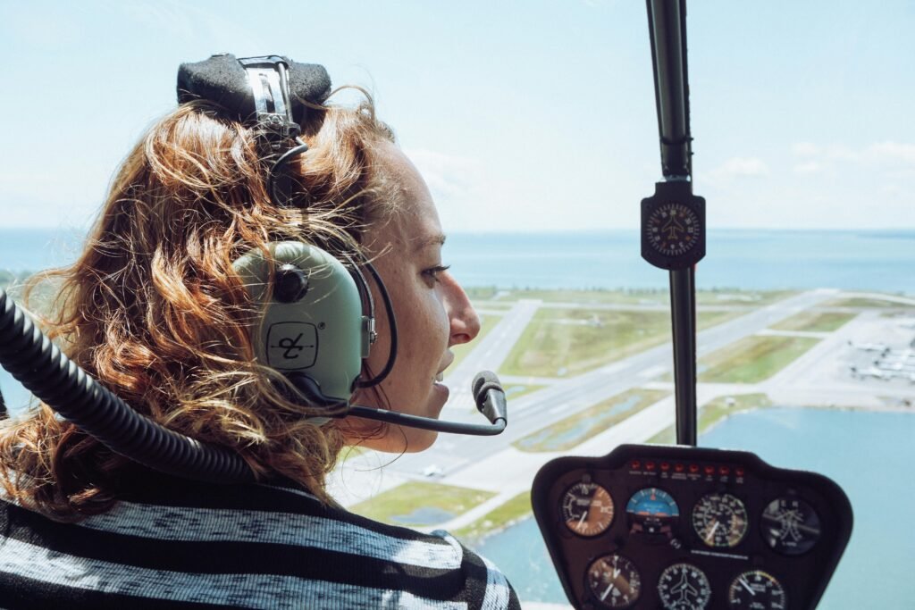 Back view of positive young female traveler in casual wear and headset sitting in cockpit of modern helicopter during flight over coastal town